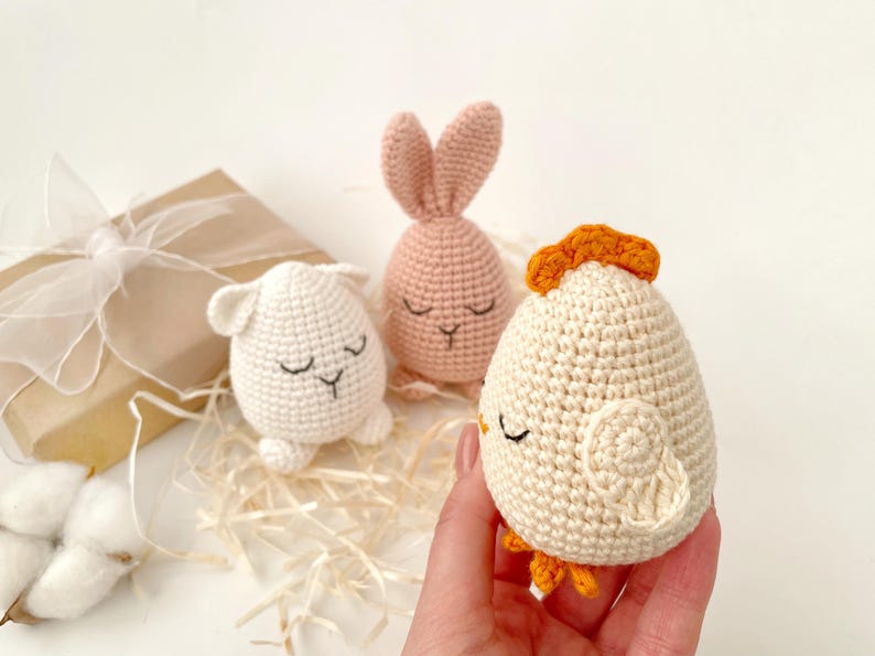 May include: Three crocheted stuffed animals shaped like an egg. The animals are a pink bunny, a white chick, and a white lamb. The animals have embroidered eyes and mouths.