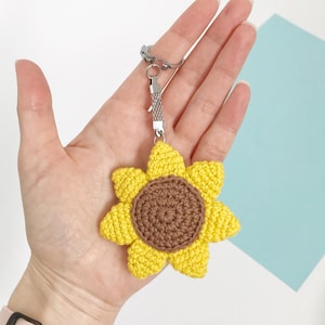 May include: A handmade crochet sunflower keychain. The sunflower has bright yellow petals and a brown center. It is attached to a silver-colored chain and clasp. The keychain is held in a person's hand.