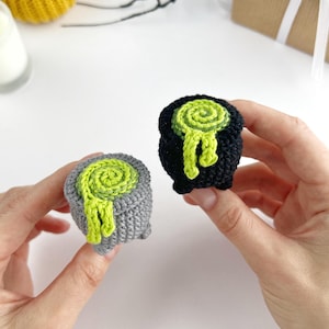 May include: Two small crocheted cauldrons, one in grey and one in black, each featuring a bright lime green spiral design on top that mimics bubbling liquid spilling over the sides. These miniature yarn pots have a textured, rounded shape.