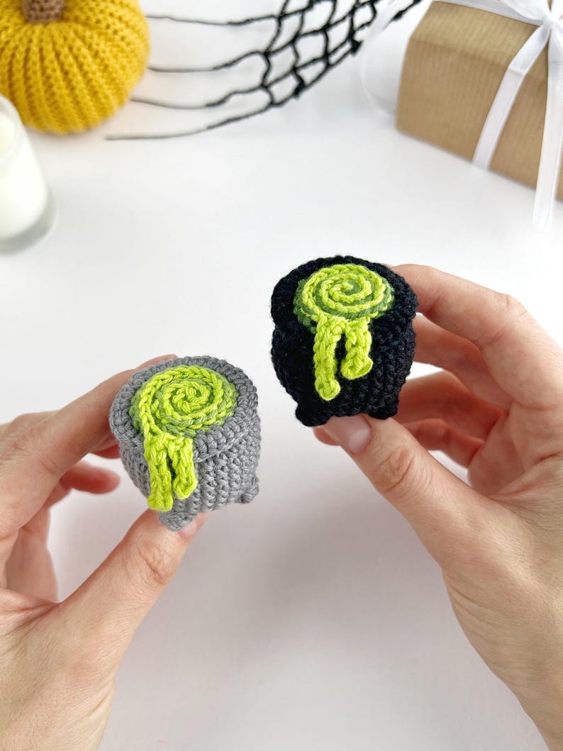 May include: Two small crocheted cauldrons, one in grey and one in black, each featuring a bright lime green spiral design on top that mimics bubbling liquid spilling over the sides. These miniature yarn pots have a textured, rounded shape.