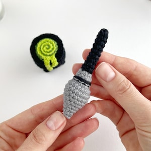 May include: A small, handcrafted crochet kunai weapon featuring a grey pointed blade and a black handle, accompanied by a separate circular black and lime green crochet scroll or target object with a spiral pattern on its top surface.