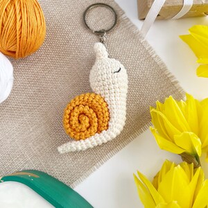 Pode incluir: Um chaveiro de caracol de croch&ecirc; com corpo branco e concha laranja. O caracol est&aacute; preso a um chaveiro prateado.