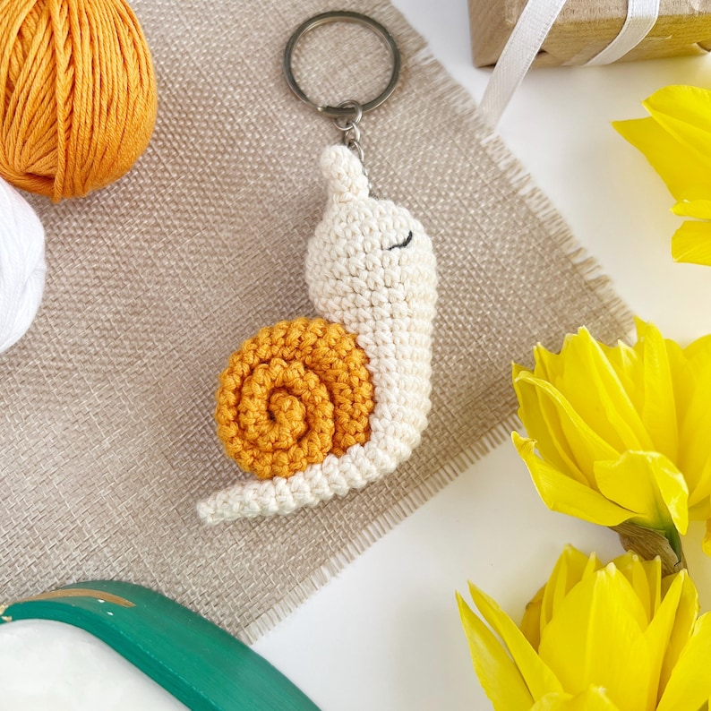 Pode incluir: Um chaveiro de caracol de croch&ecirc; com corpo branco e concha laranja. O caracol est&aacute; preso a um chaveiro prateado.