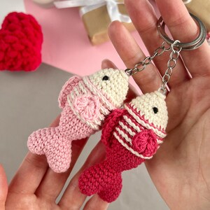 May include: Two handmade crochet fish keychains in shades of pink and red. The fish have cream-colored heads and are attached to silver keychains. The fish are detailed with textured patterns and small black eyes.
