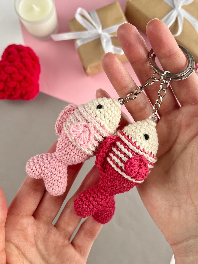 May include: Two handmade crochet fish keychains in shades of pink and red. The fish have cream-colored heads and are attached to silver keychains. The fish are detailed with textured patterns and small black eyes.