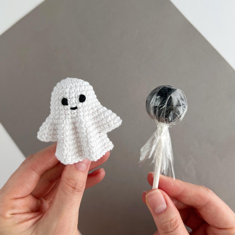 May include: A small white crocheted ghost figure with two black circular eyes and a stitched mouth, positioned next to a round black lollipop wrapped in clear plastic with a white stick, showcasing a contrast between the textile and candy.