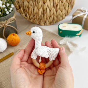 May include: A white crocheted goose rattle with an orange crocheted tail and a wooden ring. The rattle is held in a person's hands.