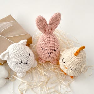 May include: Three crocheted stuffed animals: a pink bunny, a white lamb, and a white chick. The animals are all lying down with their eyes closed. They are sitting on a bed of shredded paper.