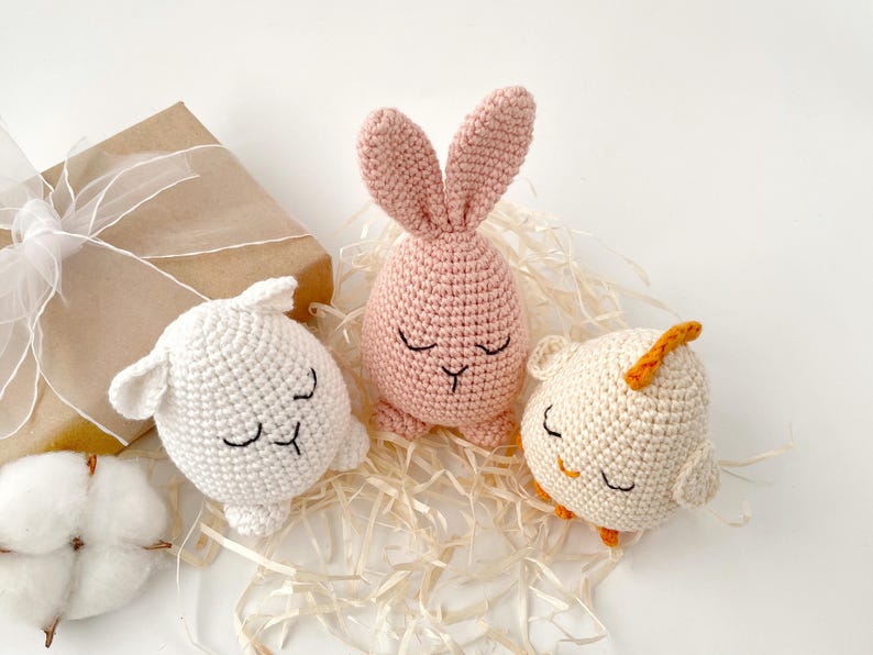 May include: Three crocheted stuffed animals: a pink bunny, a white lamb, and a white chick. The animals are all lying down with their eyes closed. They are sitting on a bed of shredded paper.