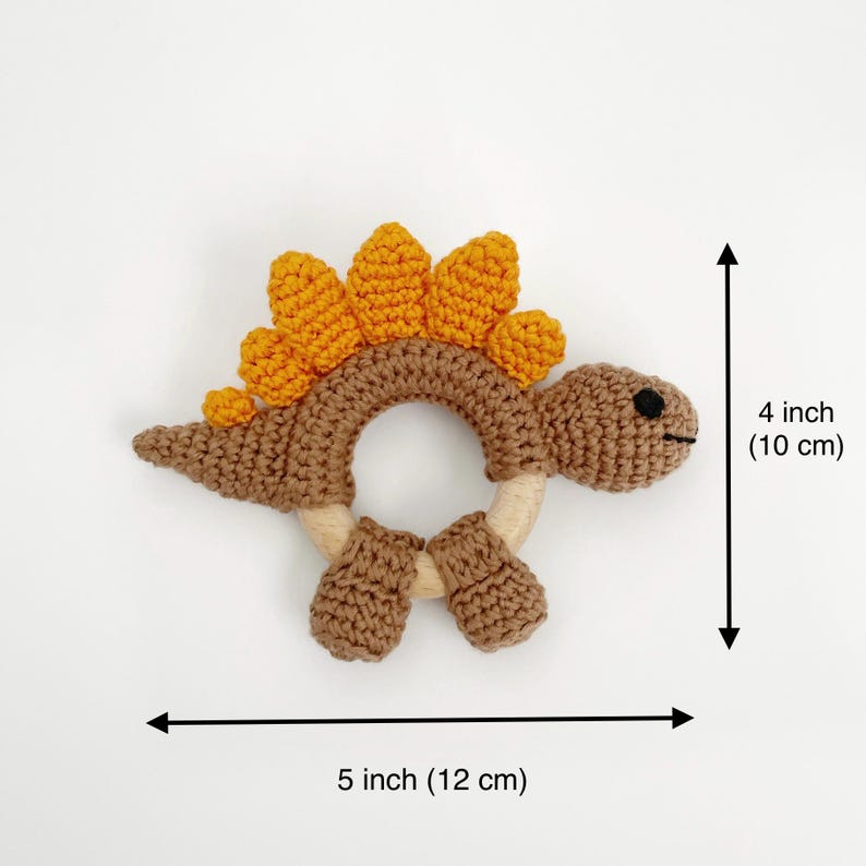 May include: A crocheted dinosaur-shaped baby teether. The toy is brown with orange spikes and a wooden ring. It measures 5 inches (12 cm) long and 4 inches (10 cm) high.