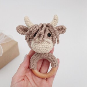 Puede incluir: Un sonajero de vaca tejido a crochet en marr&oacute;n y blanco con un anillo de madera. El sonajero se sostiene en una mano.