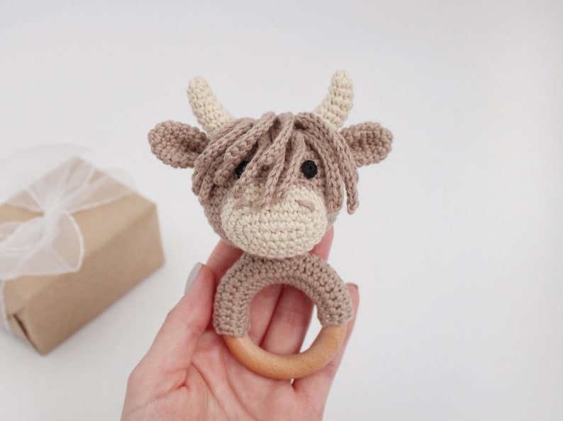 Puede incluir: Un sonajero de vaca tejido a crochet en marr&oacute;n y blanco con un anillo de madera. El sonajero se sostiene en una mano.