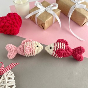 May include: Two crocheted fish in pink and red, with white accents, face each other. The fish are on a gray and pink surface, with a red heart, wrapped gifts, and a candle in the background. The fish have black eyes.
