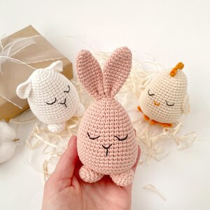 May include: Three crocheted stuffed animals: a white lamb, a pink bunny, and a white chick with orange feet. The animals have embroidered eyes and mouths.