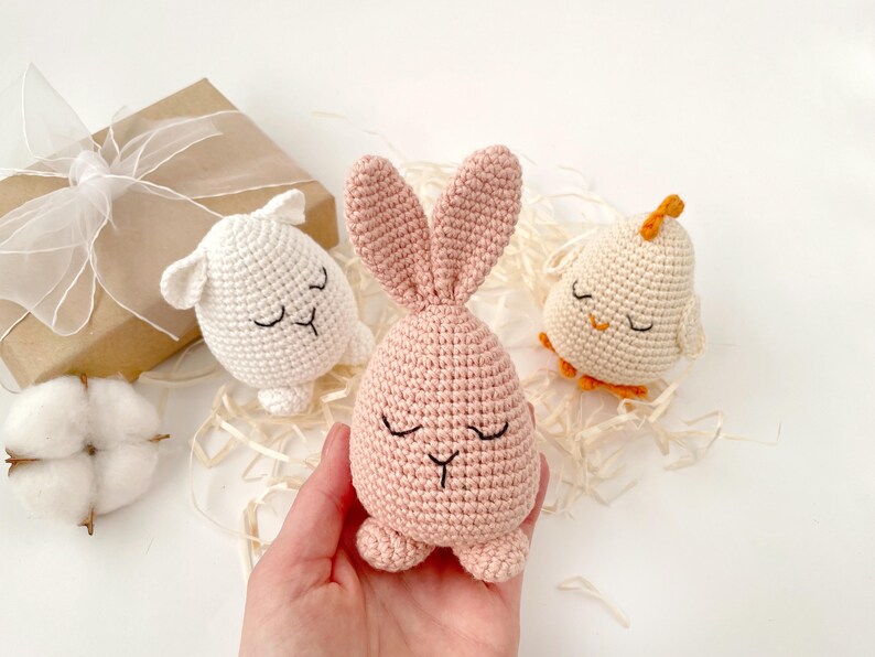May include: Three crocheted stuffed animals: a white lamb, a pink bunny, and a white chick with orange feet. The animals have embroidered eyes and mouths.