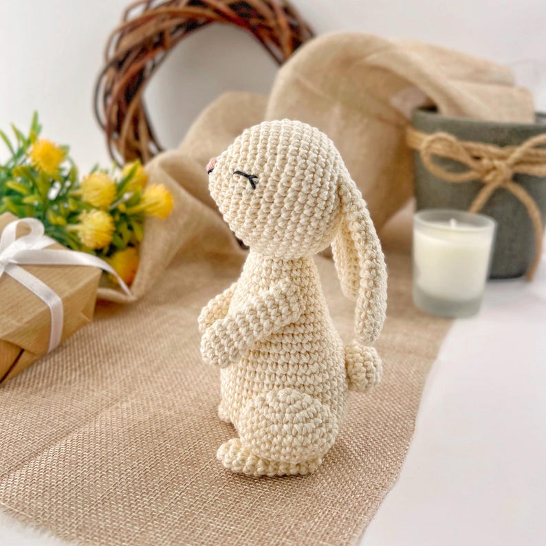 May include: A crocheted white bunny rabbit toy with closed eyes. The bunny is standing on a beige burlap surface.