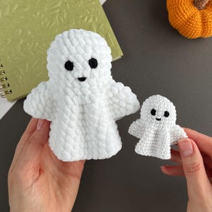 May include: Two white crochet ghost figures made of soft, plush yarn. Each ghost features two small black embroidered eyes and a tiny black mouth. The larger ghost is on the left, and the smaller ghost is on the right, both with rounded heads and arms.