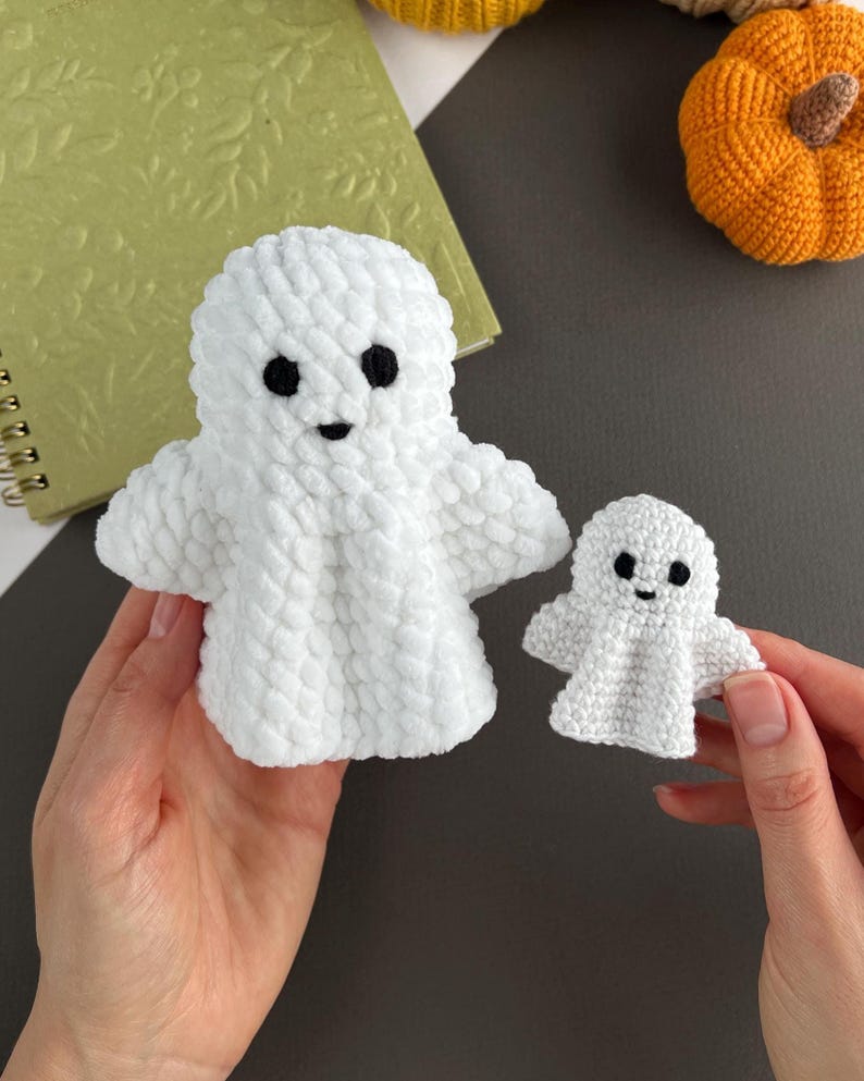 May include: Two white crochet ghost figures made of soft, plush yarn. Each ghost features two small black embroidered eyes and a tiny black mouth. The larger ghost is on the left, and the smaller ghost is on the right, both with rounded heads and arms.