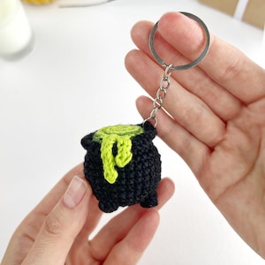 May include: A black crocheted cauldron keychain featuring a bright lime green liquid spilling over the rim. The small, round, three-legged pot is attached to a silver-toned metal split ring by a short chain, showcasing detailed yarn texture.