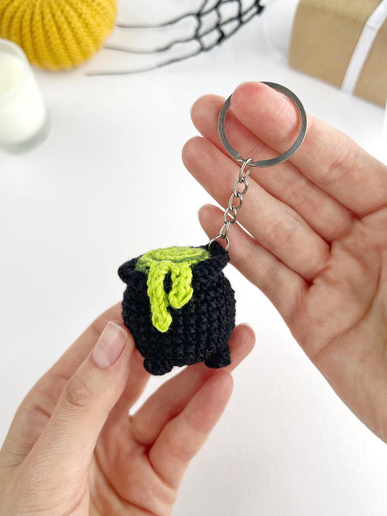 May include: A black crocheted cauldron keychain featuring a bright lime green liquid spilling over the rim. The small, round, three-legged pot is attached to a silver-toned metal split ring by a short chain, showcasing detailed yarn texture.