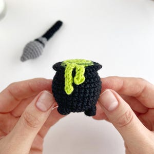 May include: A small, black crocheted cauldron with three short legs and a textured surface. A bright lime green crocheted liquid is spilling over the top rim, creating a dripping effect. The item is crafted from yarn with visible stitches.