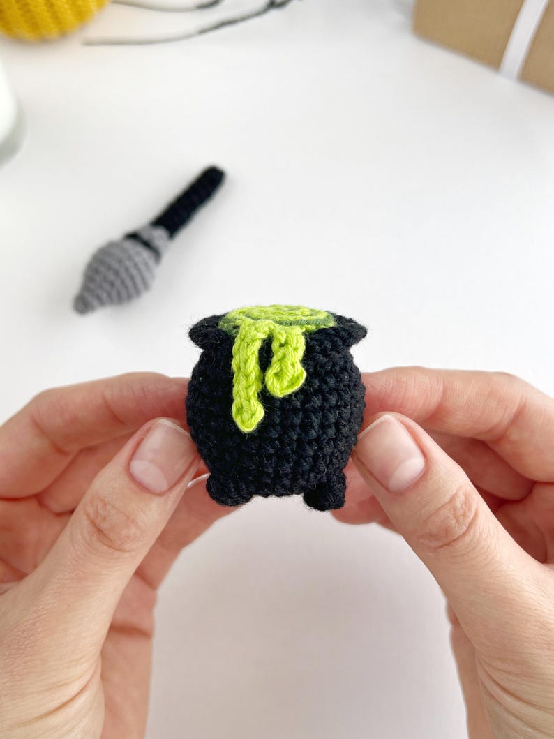 May include: A small, black crocheted cauldron with three short legs and a textured surface. A bright lime green crocheted liquid is spilling over the top rim, creating a dripping effect. The item is crafted from yarn with visible stitches.