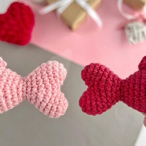 May include: Close-up of two handmade crochet bow ties, one light pink and one red. The background includes a red crochet heart, wrapped gifts with white ribbons, and a lit candle, suggesting a Valentine's Day theme.