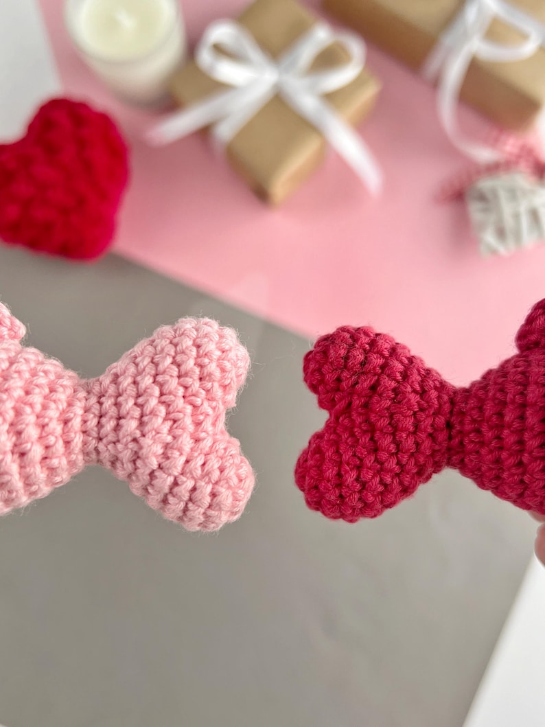 May include: Close-up of two handmade crochet bow ties, one light pink and one red. The background includes a red crochet heart, wrapped gifts with white ribbons, and a lit candle, suggesting a Valentine's Day theme.