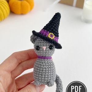 Peut inclure: Une petite figurine de chat crochetée en gris portant un chapeau de sorcière noir avec une bande violette et une boucle jaune. Le chat a des yeux de sécurité noirs, un nez brodé rose et un collier violet. Un logo noir indique PDF PATTERN IN ENGLISH.