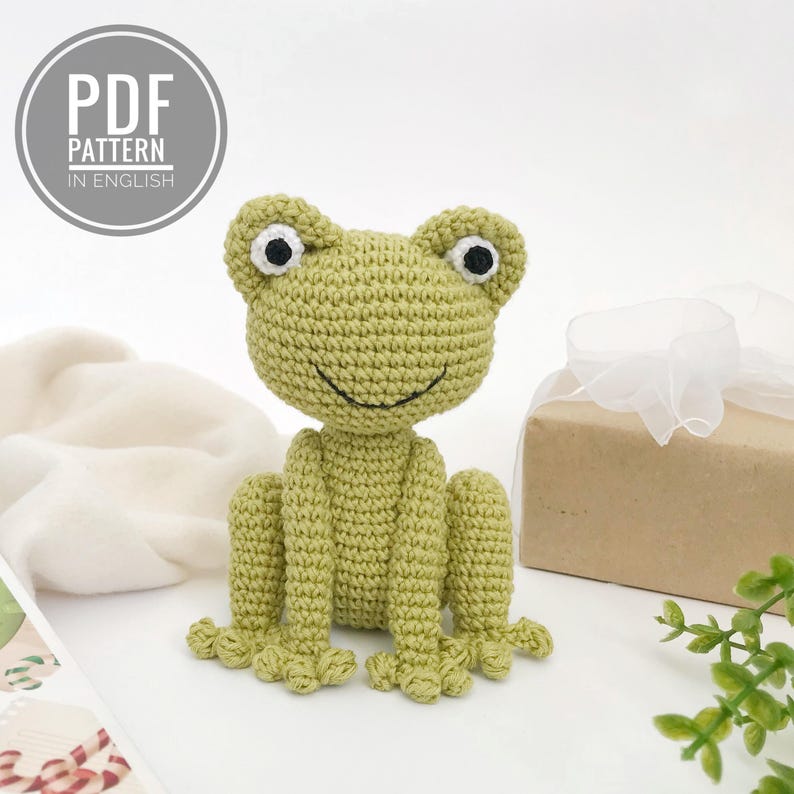 Op de afbeelding: Een gehaakte groene kikker met zwart-witte ogen en een lachende mond. De kikker zit rechtop met uitgestrekte poten. De afbeelding bevat een cadeauverpakking en de tekst "PDF PATTERN IN ENGLISH" in een cirkelvormig kader.