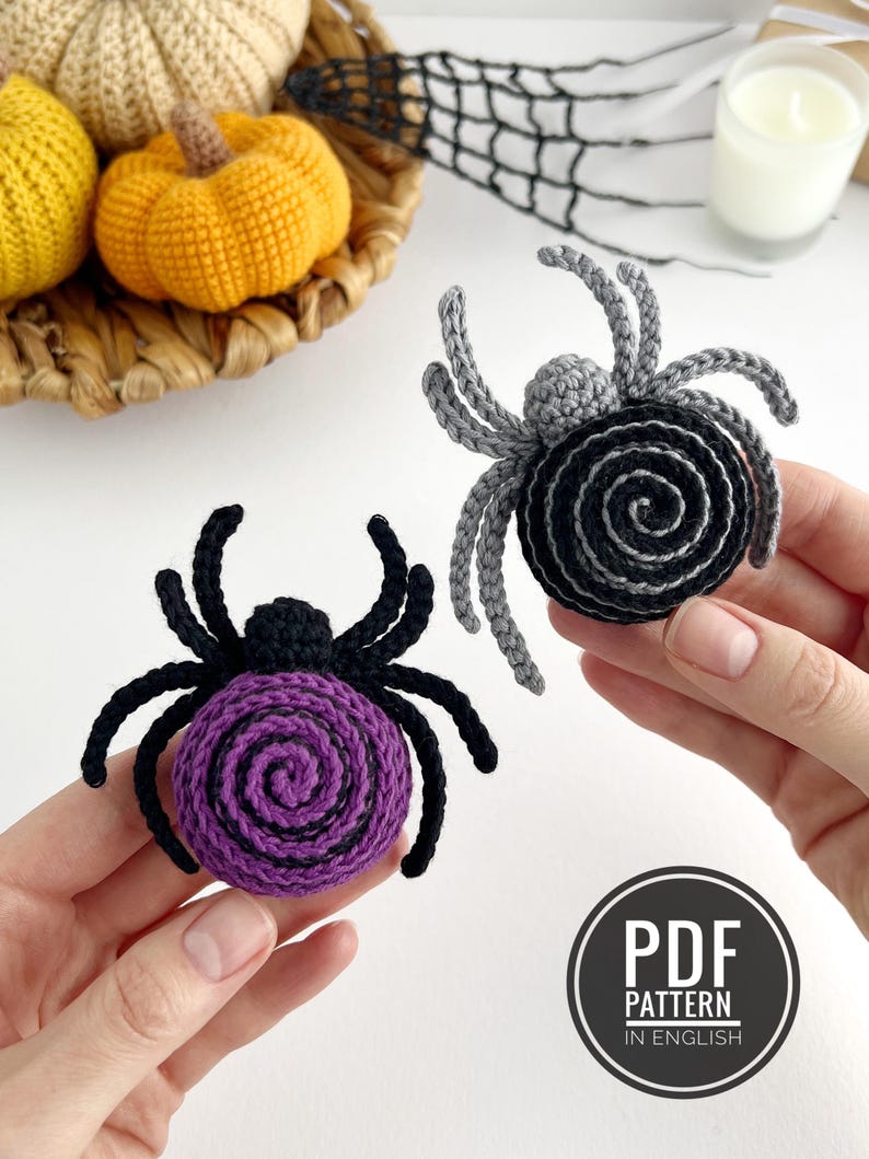 May include: Two crocheted spider ornaments, one with a purple spiral body and black legs, the other with a black and gray spiral body and gray legs. A circular black badge in the corner displays the white text PDF PATTERN IN ENGLISH.