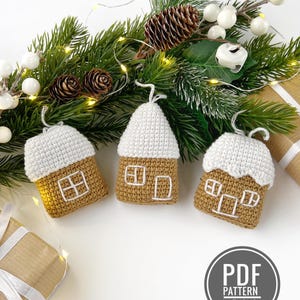 May include: Three crocheted gingerbread house ornaments featuring tan yarn walls and white yarn roofs with embroidered white windows and doors. Each house has a white yarn loop at the top for hanging. A circular badge reads PDF PATTERN IN ENGLISH.