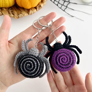 May include: Two crochet spider keychains with metal rings and chains. One spider features a black body with a gray spiral center and gray legs, while the other has a purple spiral center on a black body with black legs. Both have eight legs.