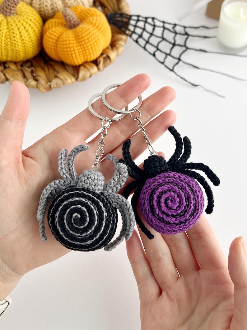 May include: Two crochet spider keychains with metal rings and chains. One spider features a black body with a gray spiral center and gray legs, while the other has a purple spiral center on a black body with black legs. Both have eight legs.