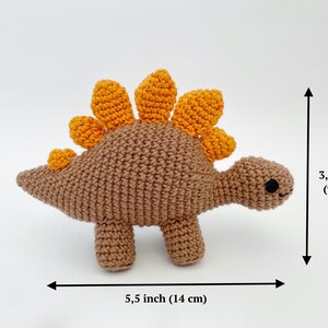 May include: A crocheted stegosaurus toy in brown with orange spines. The handmade dinosaur measures 5.5 inches (14 cm) long and 3.5 inches (9 cm) tall. The toy has black eyes and four small legs.