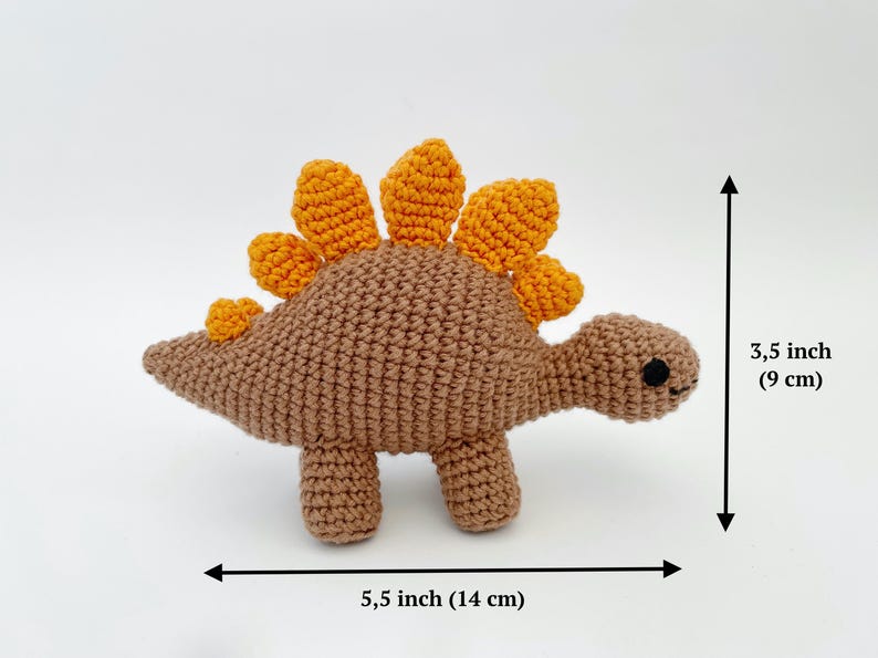 May include: A crocheted stegosaurus toy in brown with orange spines. The handmade dinosaur measures 5.5 inches (14 cm) long and 3.5 inches (9 cm) tall. The toy has black eyes and four small legs.