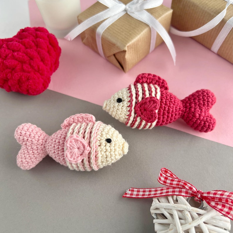 May include: Two handmade crochet fish in pink and red with white accents. One fish has a heart detail. The fish are on a gray and pink surface, with a red heart and wrapped gifts in the background. A decorative heart with a red and white ribbon is also present.