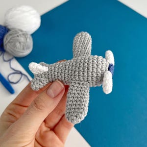 May include: A handmade gray crochet airplane with white and blue accents. The toy airplane features a propeller with white blades and a blue center. The wings and tail are also white. The background is a mix of white and blue.