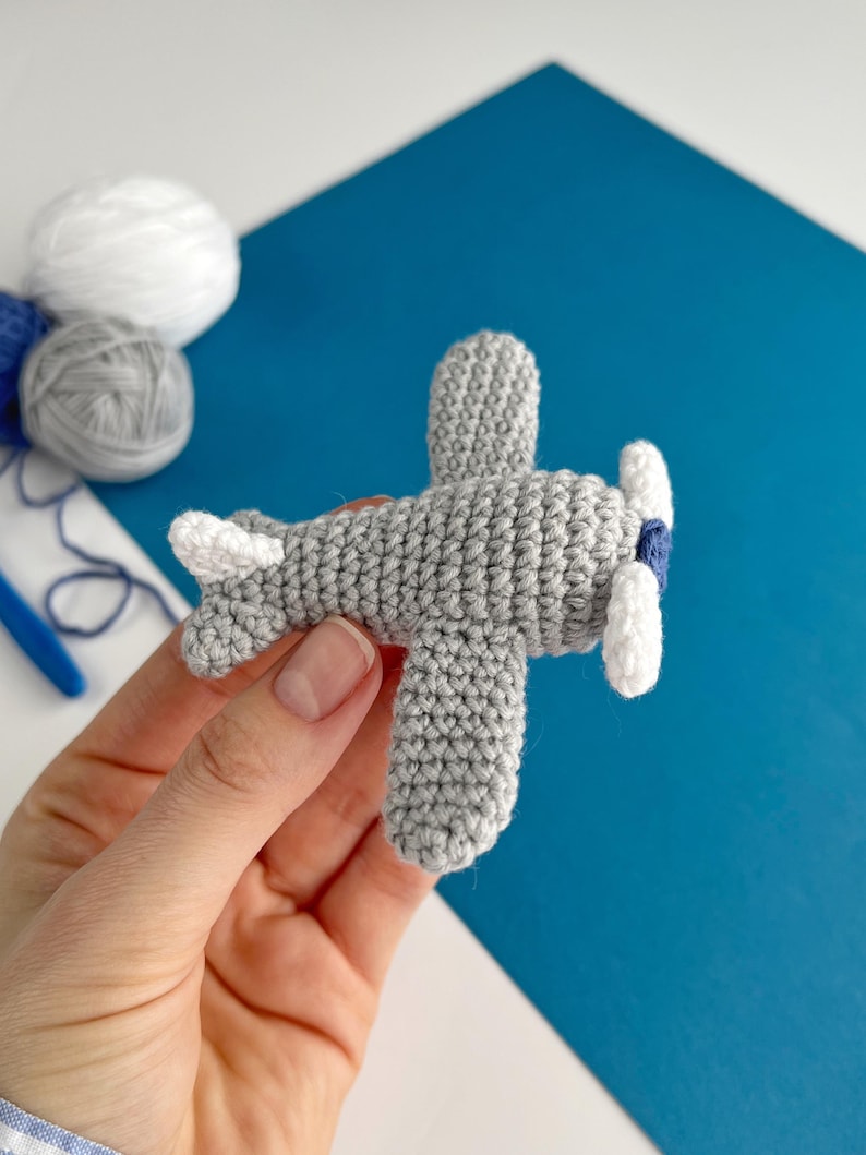 May include: A handmade gray crochet airplane with white and blue accents. The toy airplane features a propeller with white blades and a blue center. The wings and tail are also white. The background is a mix of white and blue.