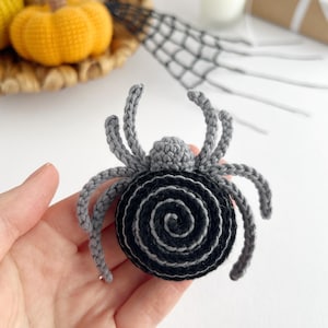 May include: A crocheted spider ornament featuring a round black and gray spiral body and eight gray legs. The spider has a small gray head section and is crafted with yarn in a detailed stitch pattern, creating a textured appearance for the arachnid.