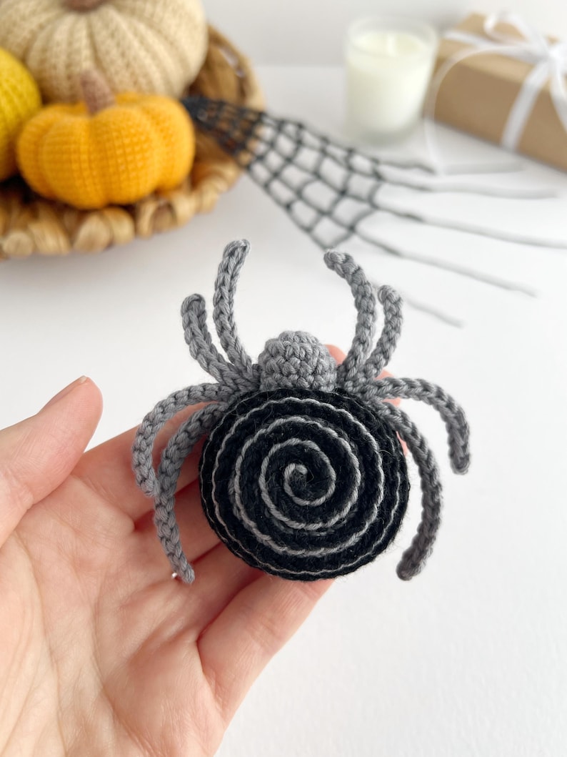 May include: A crocheted spider ornament featuring a round black and gray spiral body and eight gray legs. The spider has a small gray head section and is crafted with yarn in a detailed stitch pattern, creating a textured appearance for the arachnid.