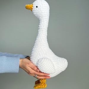 May include: A handmade white crochet goose with a long neck, yellow beak, and yellow feet. The plush toy is soft and fluffy, perfect for a nursery or as a decorative item.