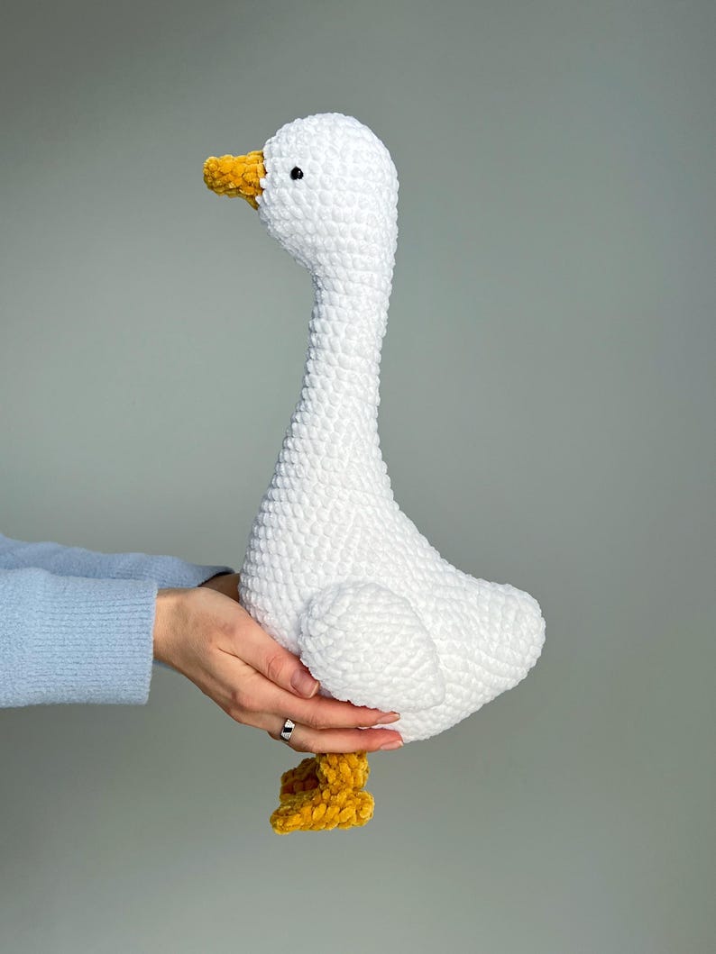 May include: A handmade white crochet goose with a long neck, yellow beak, and yellow feet. The plush toy is soft and fluffy, perfect for a nursery or as a decorative item.