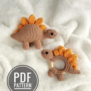 May include: Two crochet dinosaur toys in shades of brown and orange. One is a small dinosaur figure, and the other is a dinosaur-shaped rattle with a wooden ring. The toys are on a white, fluffy surface. The image includes a "PDF PATTERN" graphic.