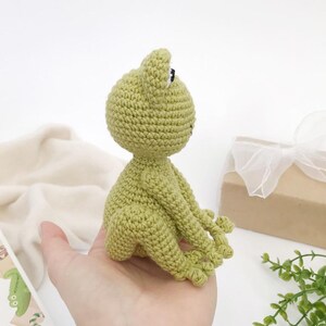 Op de afbeelding: Een handgemaakte, lichtgroene gehaakte kikkerpop met zwarte ogen en een kleine mond. De kikker zit met uitgestrekte armen en benen. Een geschenkdoos met een wit lint en een boek staan op de achtergrond.