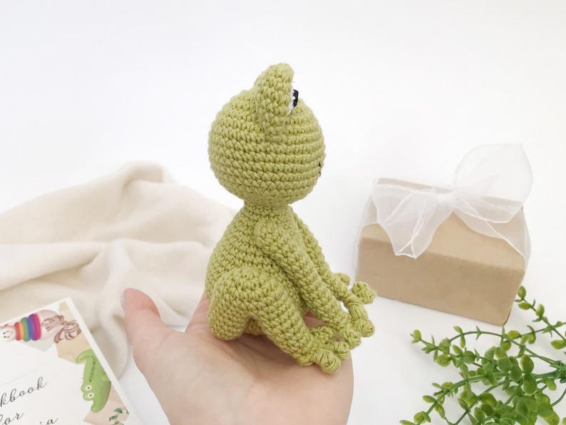Op de afbeelding: Een handgemaakte, lichtgroene gehaakte kikkerpop met zwarte ogen en een kleine mond. De kikker zit met uitgestrekte armen en benen. Een geschenkdoos met een wit lint en een boek staan op de achtergrond.