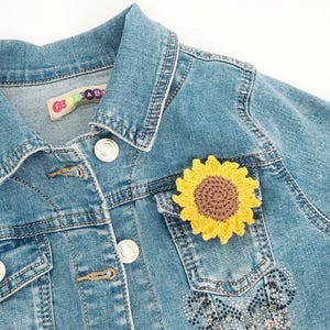 May include: A light blue denim jacket with a yellow and brown crochet sunflower patch on the pocket. The jacket has silver buttons and a small label near the collar. The jacket is a children's size.
