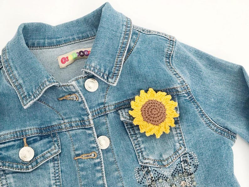 May include: A light blue denim jacket with a yellow and brown crochet sunflower patch on the pocket. The jacket has silver buttons and a small label near the collar. The jacket is a children's size.