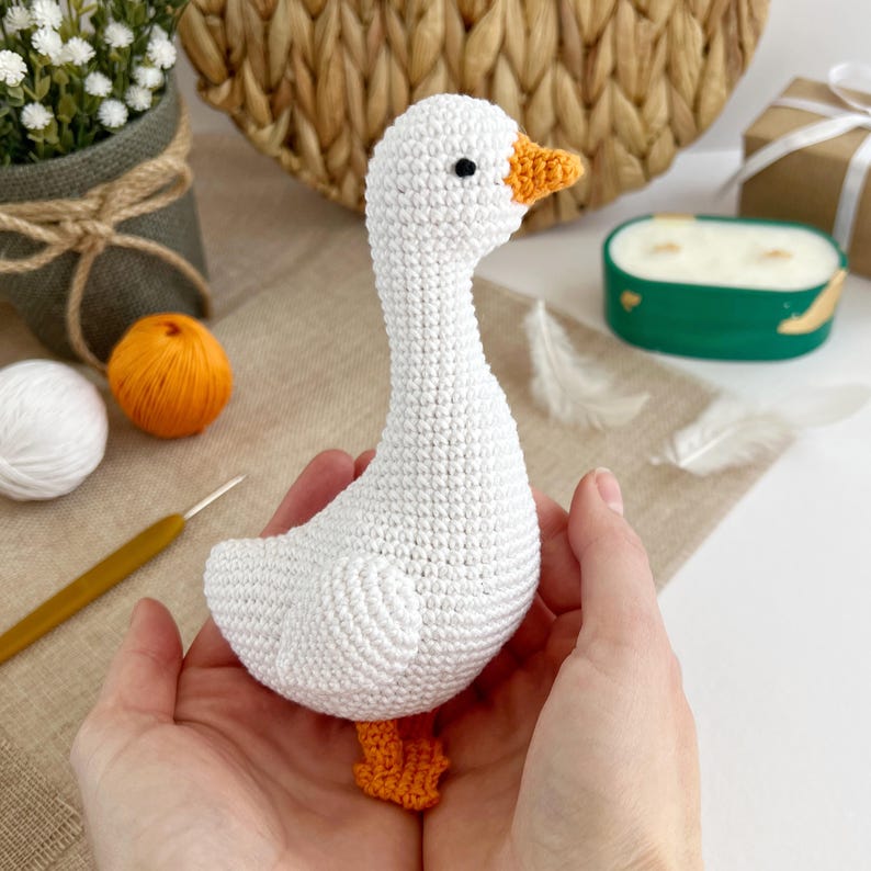 May include: A handmade white crochet goose with an orange beak and feet. The goose has a black eye and is held in open hands. The crochet goose is a decorative item.