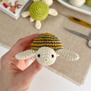 Puede incluir: Una tortuga de ganchillo hecha a mano con un caparaz&oacute;n a rayas amarillas y azul marino, cabeza y extremidades color crema y ojos negros. Otra tortuga est&aacute; en el fondo, junto con hilo y material de manualidades. Un art&iacute;culo artesanal.